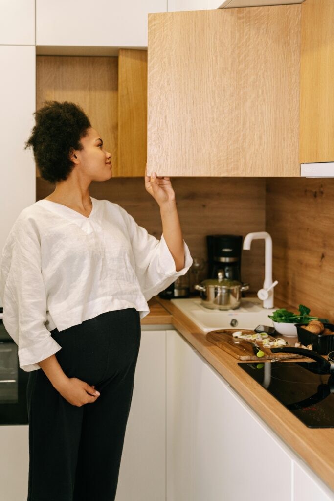 Expecting woman in a kitchen, reaching for an open cabinet, smiling warmly.